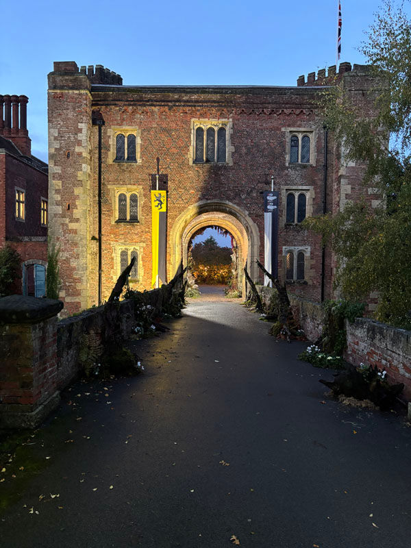 brick priory gate with arched entrance, banners and a path leading through, framed by trees under a blue sky.