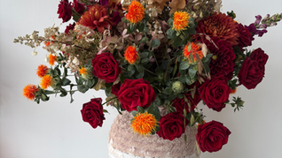 Colourful bouquet of red roses, orange flowers and dried beige foliage in a textured white vase on a white table against a plain wall