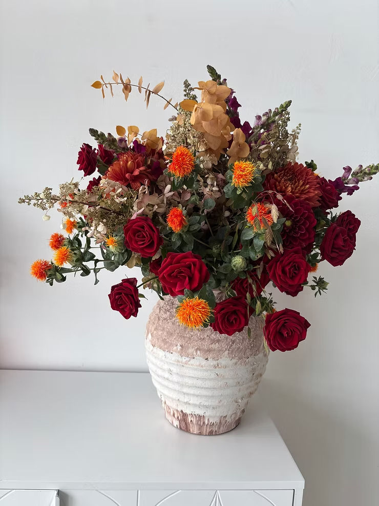Colourful bouquet of red roses, orange flowers and dried beige foliage in a textured white vase on a white table against a plain wall