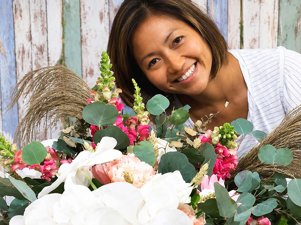 Smiling woman leaning over a colorful bouquet of white and pink flowers with greenery against a rustic painted wood backdrop