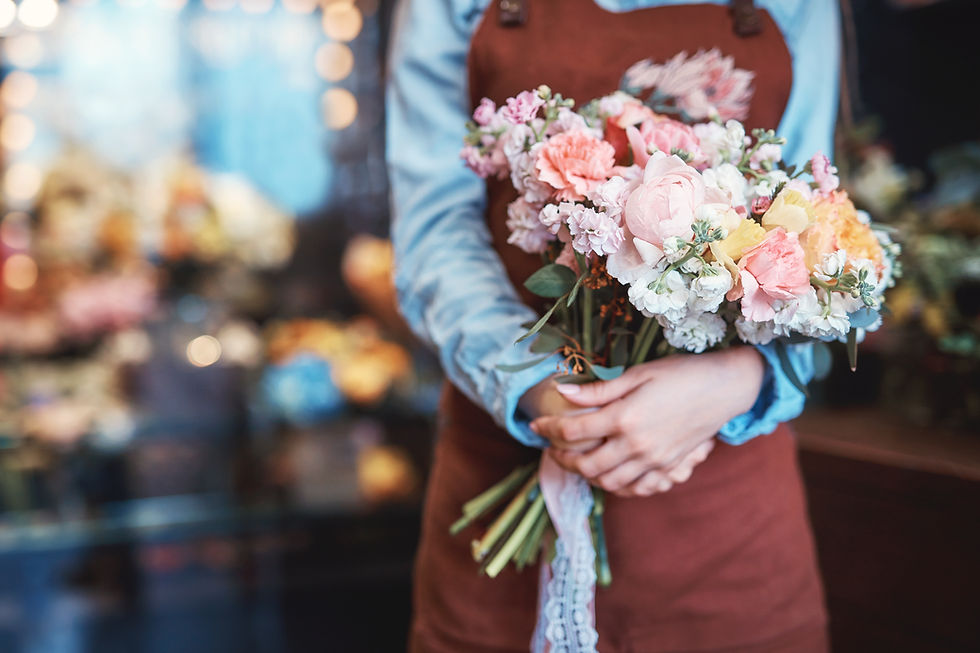 Person in a blue shirt and brown apron holds a pastel pink-and-white flower bouquet in a blurred flower shop.