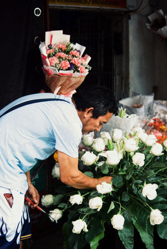 Man in a flower shop arranging white roses, with a bouquet of pink flowers over his shoulder.