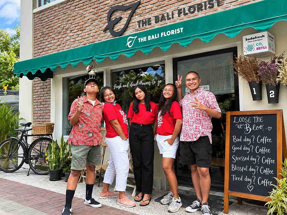 Five smiling staff pose outside The Bali Florist café under green awning; chalkboard says Coffee for any day.