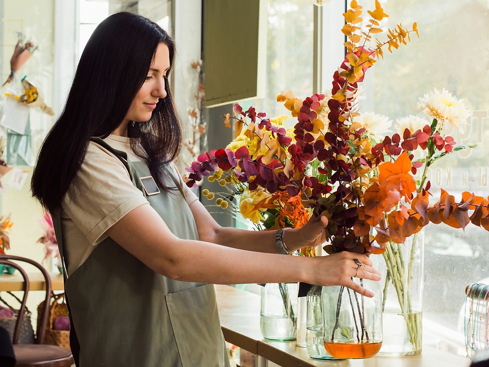 Woman in a flower shop arranging colorful bouquets by a bright window, focused and calm, with orange, red, and white blooms in vases.