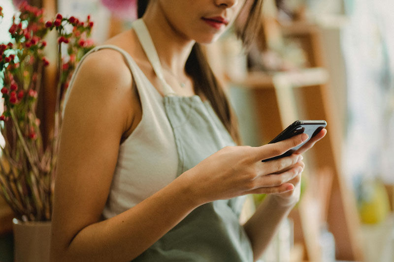 Woman in a green apron checks her smartphone indoors, with flowers and soft blurred background, focused and calm