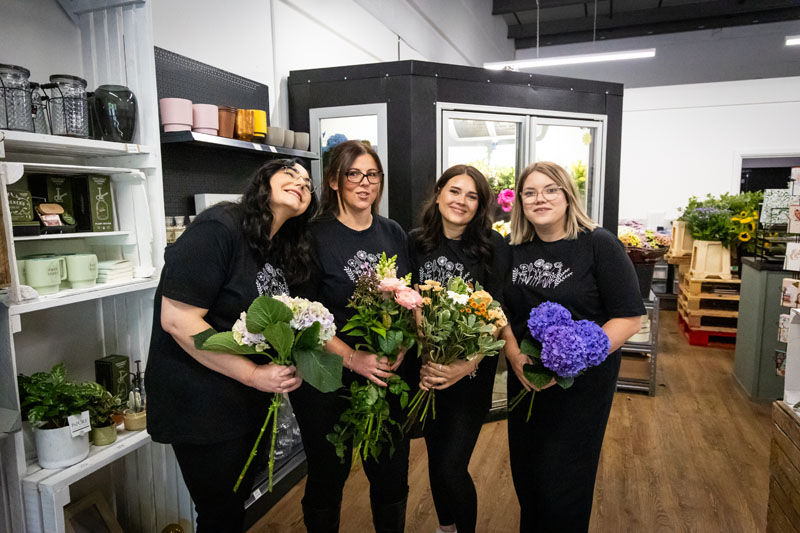 Four women in black shirts smile in a flower shop, holding colorful bouquets amid shelves and blooms.